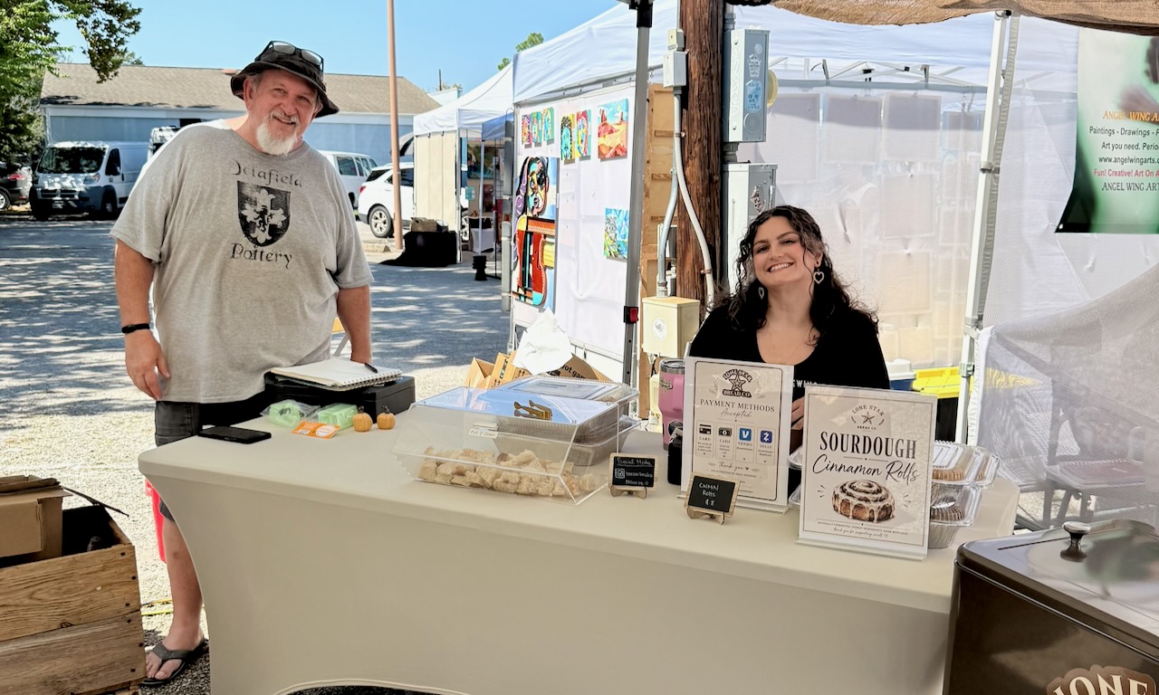 John Delafield of Delafield Pottery and Irena of Lone Star Bread Co. stand behind their shared booth table at First Saturday Arts Market, with sourdough cinnamon rolls and pottery on display under a white tent.